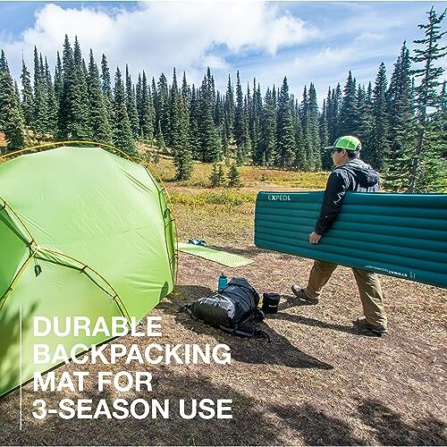 Person carrying a green backpacking mat near a tent in a forest.